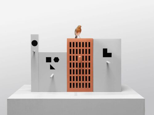 grijze vogelhuisjes op een rij met zwarte abstracte geometrische gaten met een steenrood vogelhuis in het midden waar een roodborstje op het dak zit