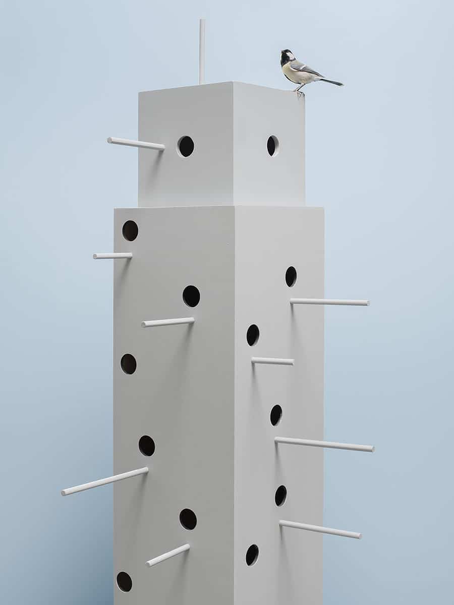 hoog vogelhuis met verschillende zwarte gaten en uitstekende stokken met op het dak met een koolmees tegen een hemelsblauwe achtergrond