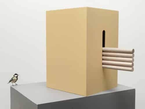 Groot okerkleurig vogelhuis op grijze sokkel met gleuf en vijf stokken op elkaar met op de hoek van de sokkel een koolmees